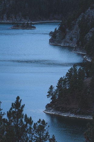 A serene lake bordered by rocky cliffs and dense pine trees, creating a tranquil and secluded atmosphere. The water is a deep blue, reflecting the surrounding nature and blending seamlessly with the dark hues of the trees and rocks.