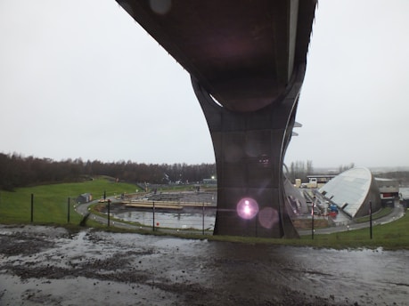 A towering structure dominates the foreground, likely supporting a bridge or overpass. Below, a fenced area contains construction materials and equipment. A rounded, modern building with a sloped roof appears to be part of an industrial or technological facility. In the background, trees line the horizon under an overcast sky, and the ground is covered with wet soil and patches of grass.