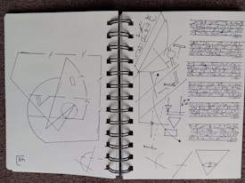 A spiral-bound notebook is open to two pages filled with detailed geometric sketches and what appears to be text written in a stylized script. On the left page, there are various geometric shapes, including circles, triangles, and intersecting lines, annotated with measurements. The right page features more geometric patterns and several lines of text, written in a unique, flowing script. The dominant colors are minimal, mostly black ink on white paper.