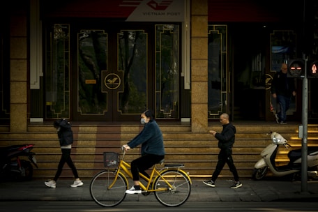 People pass by the entrance of a building with Vietnam Post signage. A person rides a yellow bicycle while wearing a mask, and another individual looks at a phone. Two people are walking, one with their hands in their pockets, and a scooter is parked nearby. Traffic lights signal the pedestrian crossing.