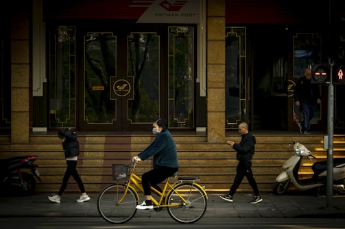 People pass by the entrance of a building with Vietnam Post signage. A person rides a yellow bicycle while wearing a mask, and another individual looks at a phone. Two people are walking, one with their hands in their pockets, and a scooter is parked nearby. Traffic lights signal the pedestrian crossing.