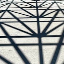 Geometric shadows create an abstract pattern on a light concrete surface, composed of intersecting lines forming triangles and angles.