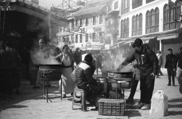 A bustling street scene features people gathered around large cooking pots with steam rising, suggesting food preparation or street food vending. The background includes several buildings with traditional architectural elements, and a crowd of people walking by, creating a lively atmosphere.