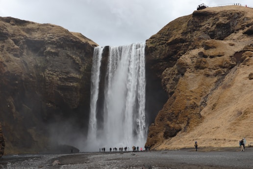 A large waterfall cascades down a rugged cliff face, surrounded by rocky terrain with sparse vegetation. At the base, a group of people stand, appearing small in comparison to the towering waterfall. Above the falls, more people are visible along a walking path.