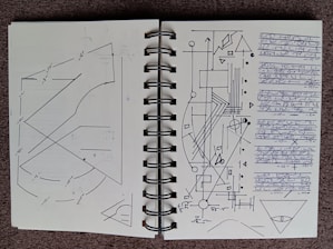 A notebook is open to a double-page spread filled with geometric diagrams, arrows, and lines. The left page features intersecting lines and curves with numerical annotations, possibly measurements. The right page contains a complex arrangement of geometric shapes, including triangles, circles, and rectangles, along with arrows and connecting lines. There is a block of indecipherable, handwritten script on the right edge. Both pages have a minimalist, technical aesthetic.