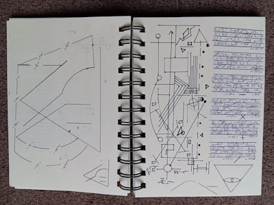 A notebook is open to a double-page spread filled with geometric diagrams, arrows, and lines. The left page features intersecting lines and curves with numerical annotations, possibly measurements. The right page contains a complex arrangement of geometric shapes, including triangles, circles, and rectangles, along with arrows and connecting lines. There is a block of indecipherable, handwritten script on the right edge. Both pages have a minimalist, technical aesthetic.