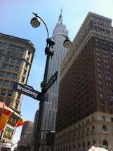 A view of towering skyscrapers including a prominent iconic building set against a clear blue sky. Street signs for Broadway and West 34th St are visible, with streetlights and a partially visible food cart adding a typical urban scene.