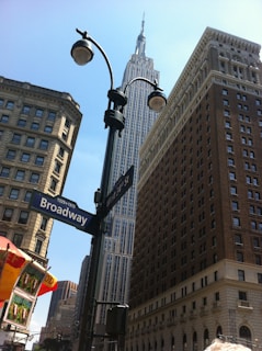 A view of towering skyscrapers including a prominent iconic building set against a clear blue sky. Street signs for Broadway and West 34th St are visible, with streetlights and a partially visible food cart adding a typical urban scene.