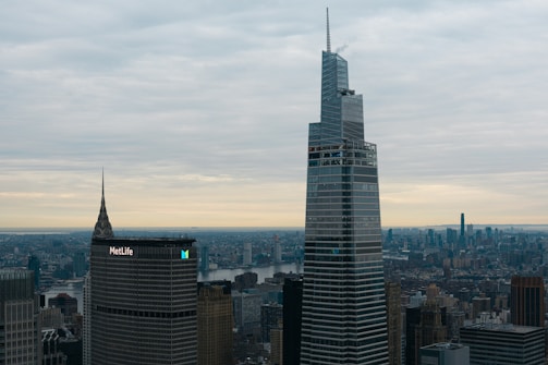 A cityscape featuring tall skyscrapers with modern architecture, set against a cloudy sky. The foreground displays prominent buildings with the MetLife building recognizable by its logo.