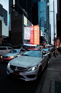 A bustling urban street scene featuring several cars, including a prominent white Mercedes. Bright digital billboards and tall skyscrapers dominate the background, suggesting a city center. The streets are busy with traffic and pedestrians can be seen on the sidewalk.
