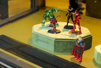 A display case featuring five superhero figurines made from crystal or glass. The figurines, representing well-known comic book characters, are posed dynamically on a green velvet pedestal. An engraved Swarovski logo is visible on the side of the pedestal. The setting is brightly lit, highlighting the vibrant colors of the red, green, and black figurines.