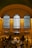 A spacious and grand hall with high arching windows, illuminated with warm lighting. The area is bustling with numerous people walking and gathering at various points. The ceiling displays intricate architectural details, and there are festive wreaths adding a holiday touch to the ambiance.