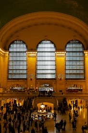 A spacious and grand hall with high arching windows, illuminated with warm lighting. The area is bustling with numerous people walking and gathering at various points. The ceiling displays intricate architectural details, and there are festive wreaths adding a holiday touch to the ambiance.