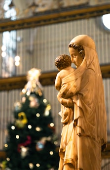 A statue depicts a serene woman in flowing robes holding a child, set against a softly lit background. The scene is peaceful with a focus on motherly affection. In the background, there appears to be a decorated Christmas tree with blurred lights creating a bokeh effect.