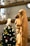 A statue depicts a serene woman in flowing robes holding a child, set against a softly lit background. The scene is peaceful with a focus on motherly affection. In the background, there appears to be a decorated Christmas tree with blurred lights creating a bokeh effect.