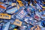 A collection of New York-themed souvenirs and memorabilia, including license plates, keychains, and magnets. Many items feature the words 'New York' and iconic imagery such as the Statue of Liberty and the American flag. The scene is dense with items overlapping each other, creating a cluttered and busy visual impression.