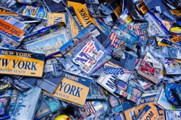 A collection of New York-themed souvenirs and memorabilia, including license plates, keychains, and magnets. Many items feature the words 'New York' and iconic imagery such as the Statue of Liberty and the American flag. The scene is dense with items overlapping each other, creating a cluttered and busy visual impression.