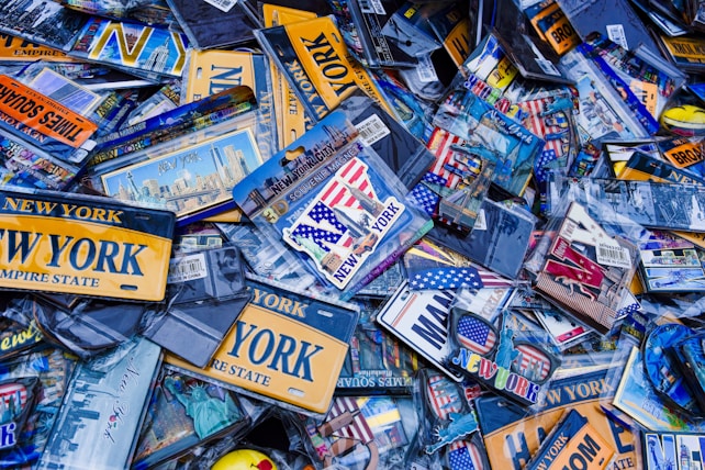 A collection of New York-themed souvenirs and memorabilia, including license plates, keychains, and magnets. Many items feature the words 'New York' and iconic imagery such as the Statue of Liberty and the American flag. The scene is dense with items overlapping each other, creating a cluttered and busy visual impression.