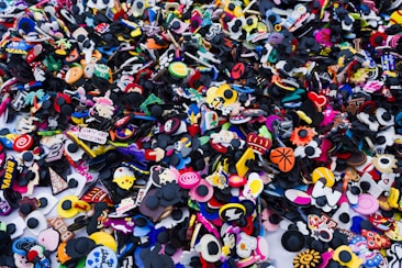 A large collection of variously colored and themed shoe charms scattered together. These charms are made of rubber and feature diverse designs including cartoon characters, symbols, and objects like basketballs and hearts. The pile creates a vibrant and chaotic visual pattern with no specific order.