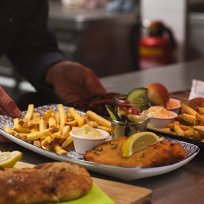 A plate of delicious food featuring golden french fries, a piece of fried fish with a lemon wedge, small bowls of sauce, and a fresh salad garnished with cucumber and cherry tomatoes. The setting appears to be a kitchen or restaurant with a hand placing the dish on a counter.