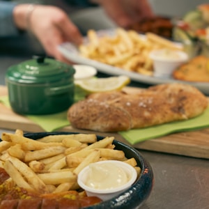 Various items of food including a bowl of fries with sauce, a small green pot, a piece of bread on a plate, and someone's hand holding a dish.