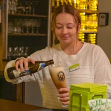 A person in a cozy bar setting is pouring a light-colored beer from a bottle into a glass, creating a foamy head. The person is wearing a white sweater and a name badge, with shelves of glasses and bottles softly illuminated in the background.