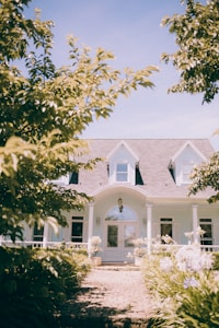 A well-maintained house with a gray gabled roof and white walls is seen at the end of a stone pathway. The front entrance has a large glass double door flanked by tall windows and decorative plants. Surrounding the pathway, lush green foliage and flowering plants frame the view, and a light blue sky is visible in the background.