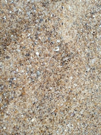 A close-up view of coarse sand consisting of a variety of small stones and pebbles. The individual particles range in color from light beige to darker shades of gray and brown.