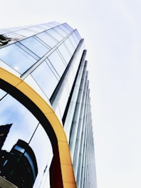 A towering modern glass building with reflective windows and a sleek design. The structure features vertical cylindrical columns alongside rows of large glass panes. There is a yellow arch framing part of the entrance, and the reflections in the glass show other buildings and a crane.