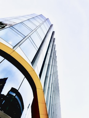 A towering modern glass building with reflective windows and a sleek design. The structure features vertical cylindrical columns alongside rows of large glass panes. There is a yellow arch framing part of the entrance, and the reflections in the glass show other buildings and a crane.