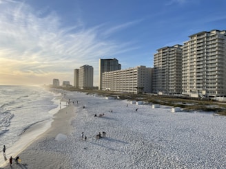 Tall beachfront buildings line the right side while a sandy beach stretches along the left, meeting the calm ocean. A few people are scattered across the beach, enjoying the open space. The sky is mostly clear with a few wispy clouds and the sun appears to be setting, casting a golden hue over the scene.