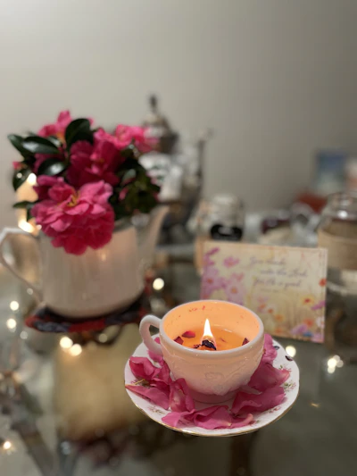 A softly lit vintage teacup candle surrounded by delicate pink peony blossoms on a cream lace tablecloth.
