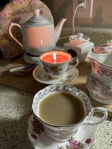 A cozy tea setup featuring an elegant white teapot, a teacup with intricate floral designs filled with tea, and another teacup holding a lit candle. There's a decorative container with a lid, a sugar bowl, and a spoon nearby. The background shows a floral pattern cushion and a wooden board, all resting on a speckled countertop.