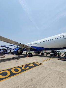 A large passenger airplane is parked on the tarmac at an airport. The aircraft has a sleek white and blue design with its airline branding visible. Ground crew members are visible nearby, working and managing logistics. The sky is clear, contributing to a bright and sunny atmosphere.