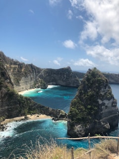 A breathtaking coastal scene with towering cliffs, turquoise waters, and a sandy beach nestled in a cove. Lush greenery covers parts of the cliffs, and the sky is bright with scattered clouds.