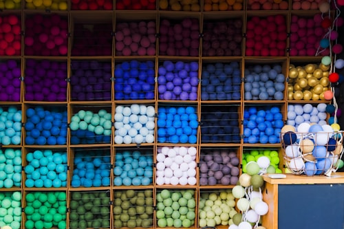 A display of shelves filled with neatly arranged colorful yarn balls in a variety of vibrant and pastel hues. Each shelf contains balls of a single color, creating a visually pleasing rainbow-like gradient. A basket filled with more yarn balls sits on a nearby table, complementing the organized array.