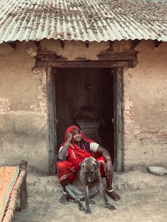 An elderly person wearing a red sari sits beside a large dog in front of a rustic, traditional mud house. The person appears contemplative or pensive, with a hand resting on their head. A charpoy, or traditional woven bed, is visible to the side.