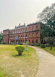 A large, historic red-brick building with multiple windows, framed by decorative arches and a sign reading 'Department of Chemistry' at the top. The structure is surrounded by a well-maintained lawn with bushes and a path leading to the entrance. Tall trees are visible on one side, adding a touch of greenery to the scene.