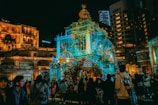 A lit-up structure decorated with colorful flowers is the focal point amidst a night-time scene against a backdrop of tall buildings. People are gathered in the area, appearing to enjoy a public event or festival.