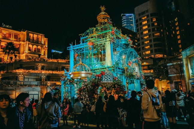 A lit-up structure decorated with colorful flowers is the focal point amidst a night-time scene against a backdrop of tall buildings. People are gathered in the area, appearing to enjoy a public event or festival.
