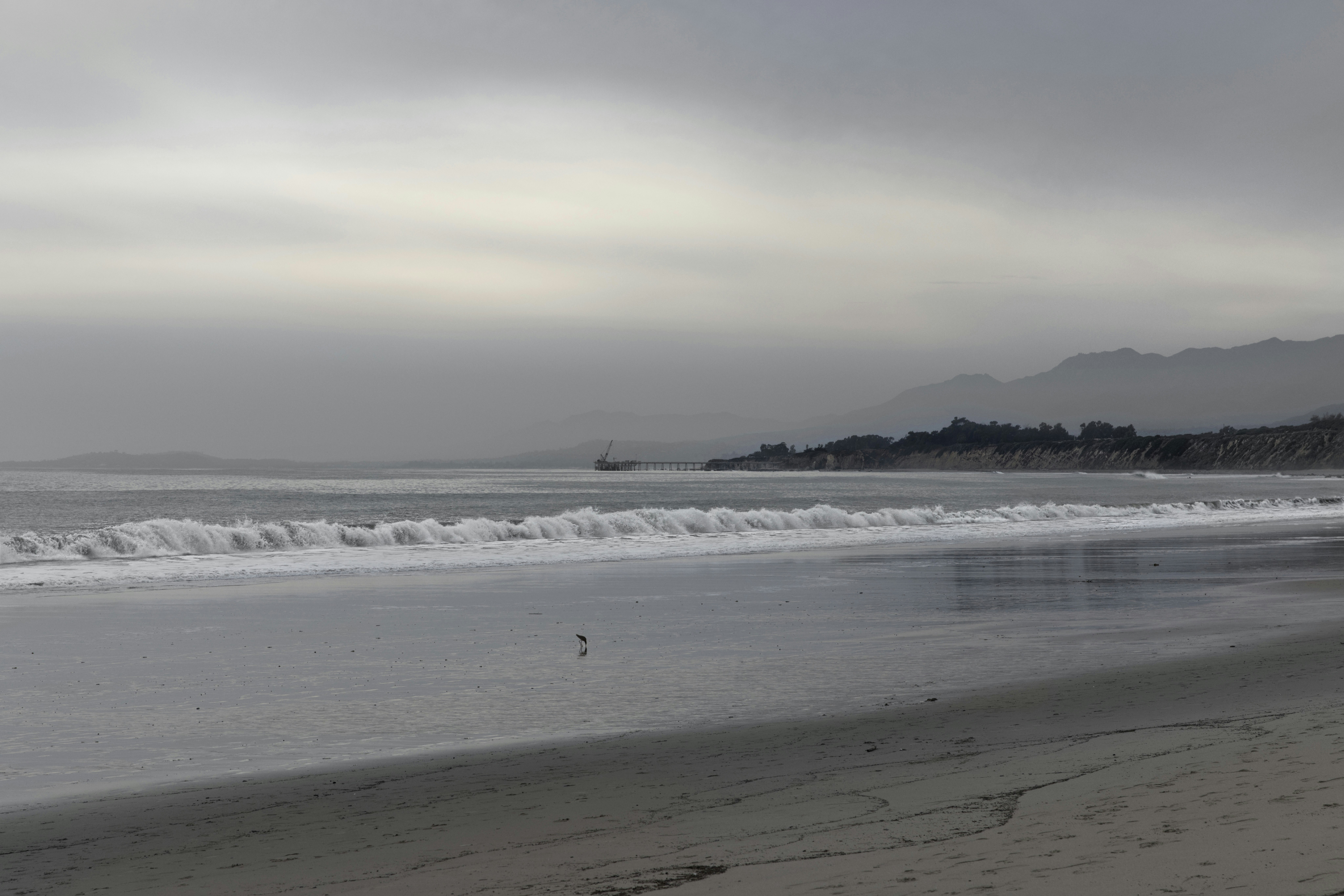 a person walking on a beach near the ocean, Cloudy skies light up Rincon Beach in Carpenteria, California with a diffuse glow.
