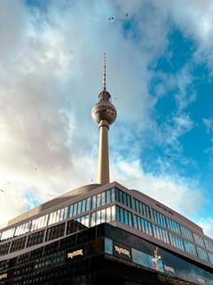 A distinctive television tower rises against a background of cloudy and blue skies. The structure's metallic sphere and antenna stand tall above a modern building with reflective glass windows. Several birds fly in the distance, adding a dynamic element to the composition.