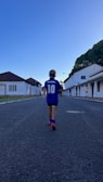 A child wearing a blue sports jersey with the name 'Maradona' and the number 10 walks down a deserted street lined with white buildings. The sky is clear and blue, and trees can be seen to the right.