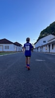 A child wearing a blue sports jersey with the name 'Maradona' and the number 10 walks down a deserted street lined with white buildings. The sky is clear and blue, and trees can be seen to the right.