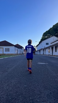 A child wearing a blue sports jersey with the name 'Maradona' and the number 10 walks down a deserted street lined with white buildings. The sky is clear and blue, and trees can be seen to the right.