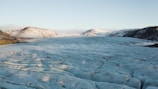 A vast glacial landscape stretches across the image, with a large expanse of ice in the foreground and rugged mountains in the background. The surface of the glacier appears to have a bluish hue with visible striations and crevices, while the mountains are partially covered with snow. The sky above is clear.