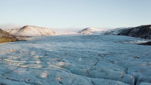 A vast glacial landscape stretches across the image, with a large expanse of ice in the foreground and rugged mountains in the background. The surface of the glacier appears to have a bluish hue with visible striations and crevices, while the mountains are partially covered with snow. The sky above is clear.