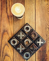 A cup of coffee with latte art sits on a wooden table next to a rustic, metal and wood tic-tac-toe game. The table has a rich grain pattern, adding warmth to the composition.
