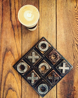 A cup of coffee with latte art sits on a wooden table next to a rustic, metal and wood tic-tac-toe game. The table has a rich grain pattern, adding warmth to the composition.
