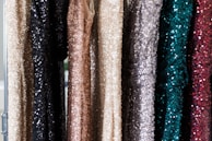 A series of sequin-covered dresses hanging on a rack, displaying a variety of colors including black, beige, gold, silver, teal, and dark red. The sequins create a shimmering effect as they catch the light.